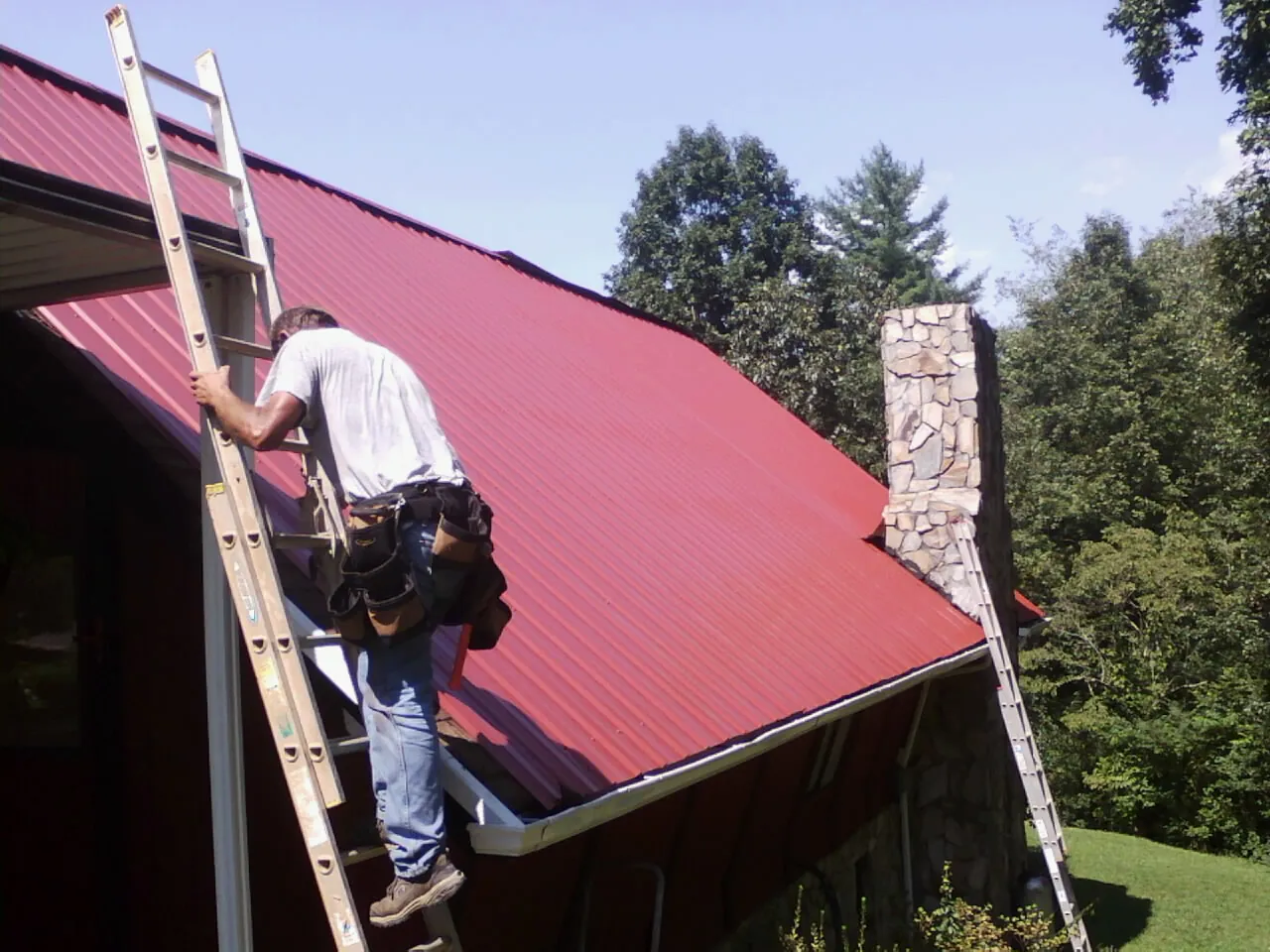 Roofer on a ladder beside a red metal roof and stone chimney — Soffit & Fascia Repair by Roofing Brothers in Gloverville
